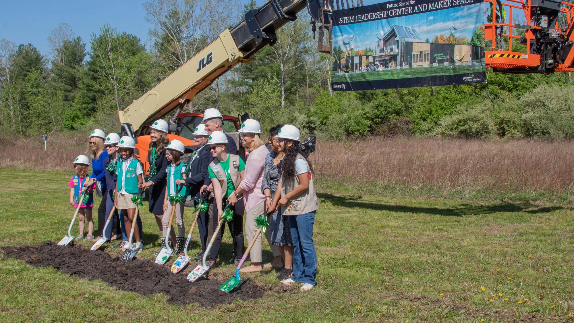 Girl Scouts and adult leaders line up with shovels that kids painted with fun pictures, for a ceremony to dig up the first bits of ground at what would become the construction site for the STEM center. There are five kids and seven adults, and they pose for a photo being taken by someone who is not shown. A big piece of construction equipment is parked behind them. 