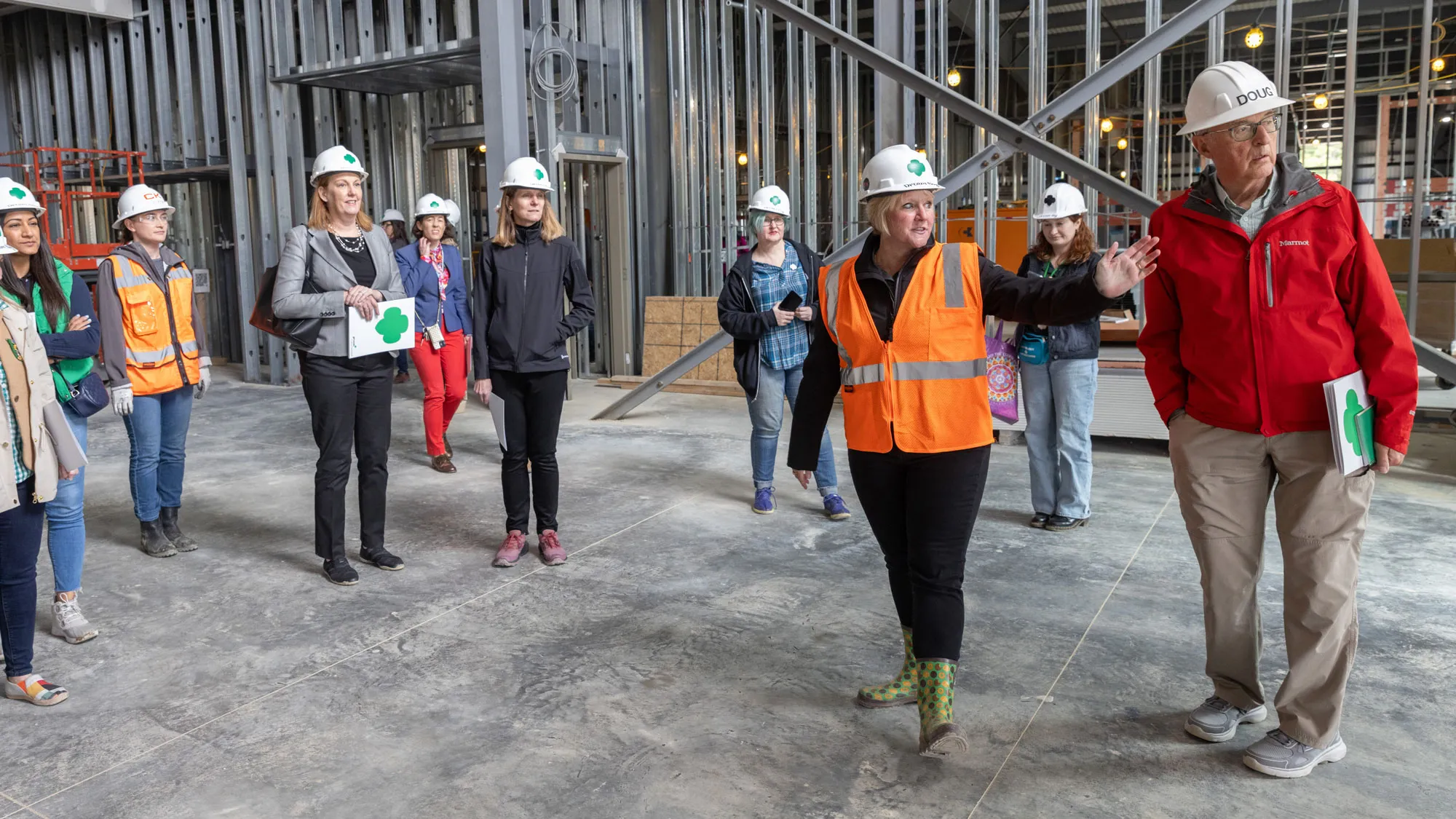 Wearing a bright construction vest and hard hat, Wharton gestures to indicate something she’s looking at off camera. The women behind her listen, many smiling, and the older man beside her looks on with rapt attention. Behind the group is the steel framework for what will be walls of a building. The concrete floor is dirty with dried mud from dozens of footprints. 