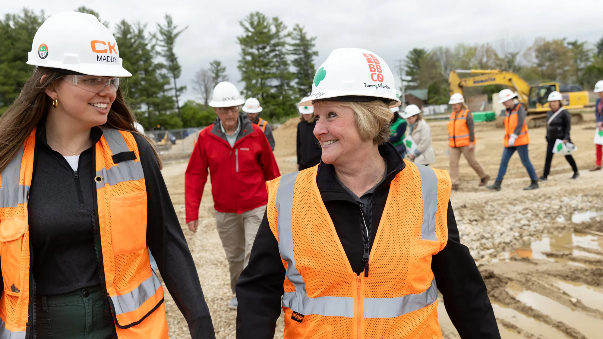 Outside at the construction site the group trails behind Wharton and a young woman who works with the construction team. They’re looking at each other and grinning, as if one has just made a joke. All people have on hard hats and safety vests. 