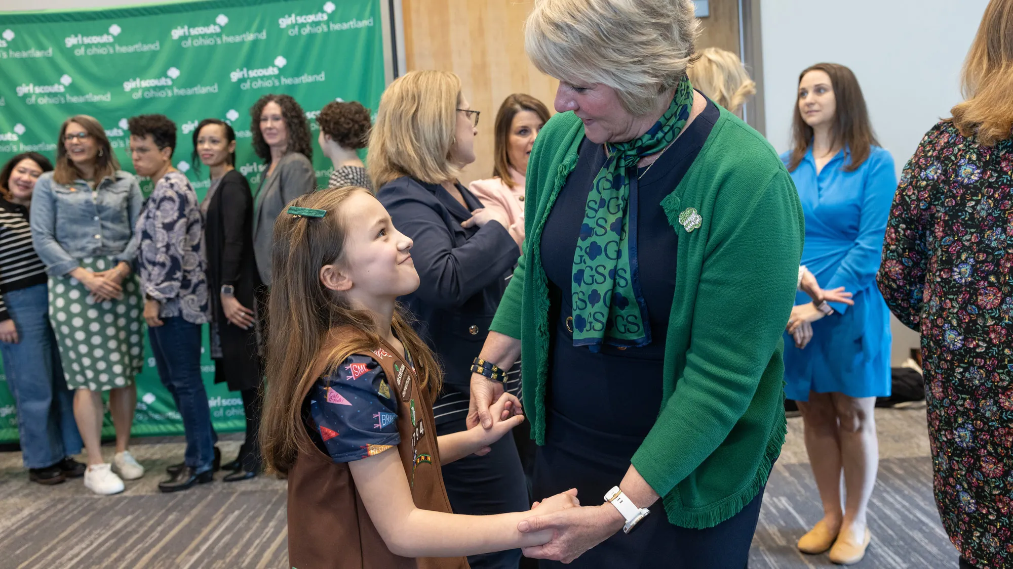 As a line of people pose for photos at the event’s commemorative photo setup behind them, Wharton clasps hands with a young girl scout, who wears her Girl Scout vest and looks up at Wharton with a mischievous smile. 