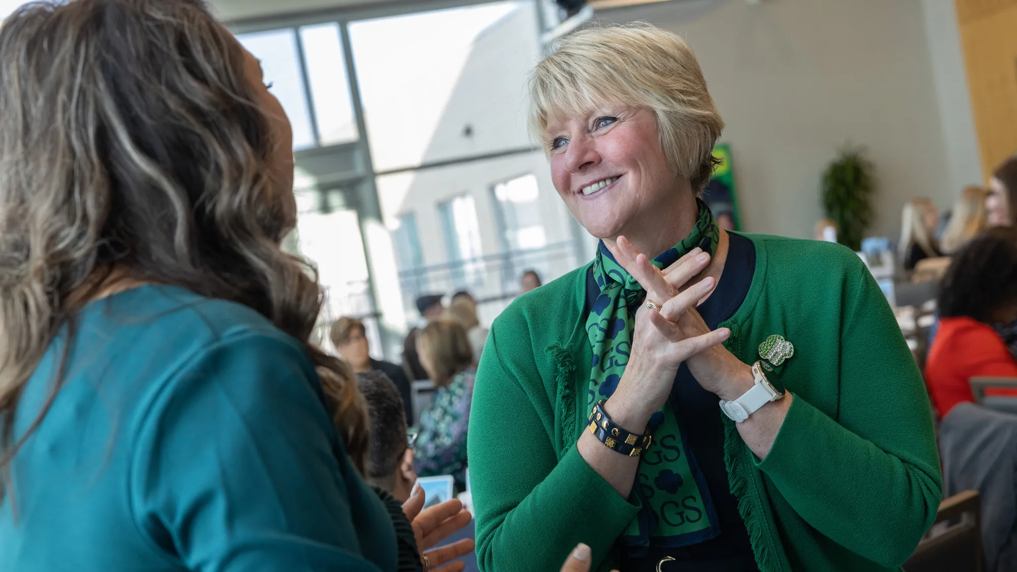 From this photo, you can tell that Tammy Wharton, a white woman with short blond hair and a pretty smile, has excellent listening habits. She seems super-engaged and even encouraging as she looks directly at the woman who’s speaking with her, who is pictured from behind. Other people are in the background of the photo, so you can see it’s a meeting or convention at a place where huge windows let in lots of natural light. 