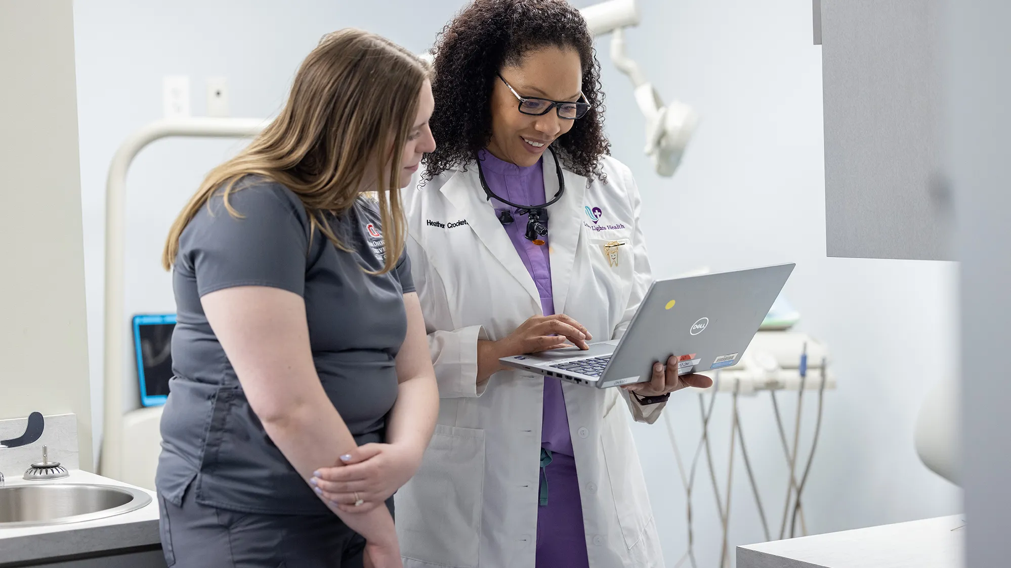 A student in Ohio State scrubs leans in to see the laptop held by a professional dentist wearing a white doctor’s coat. The dentist is a black woman with curly hair and glasses. She’s smiling as she shows the student the screen. The student is a young white woman with long hair and she seems thoughtful.