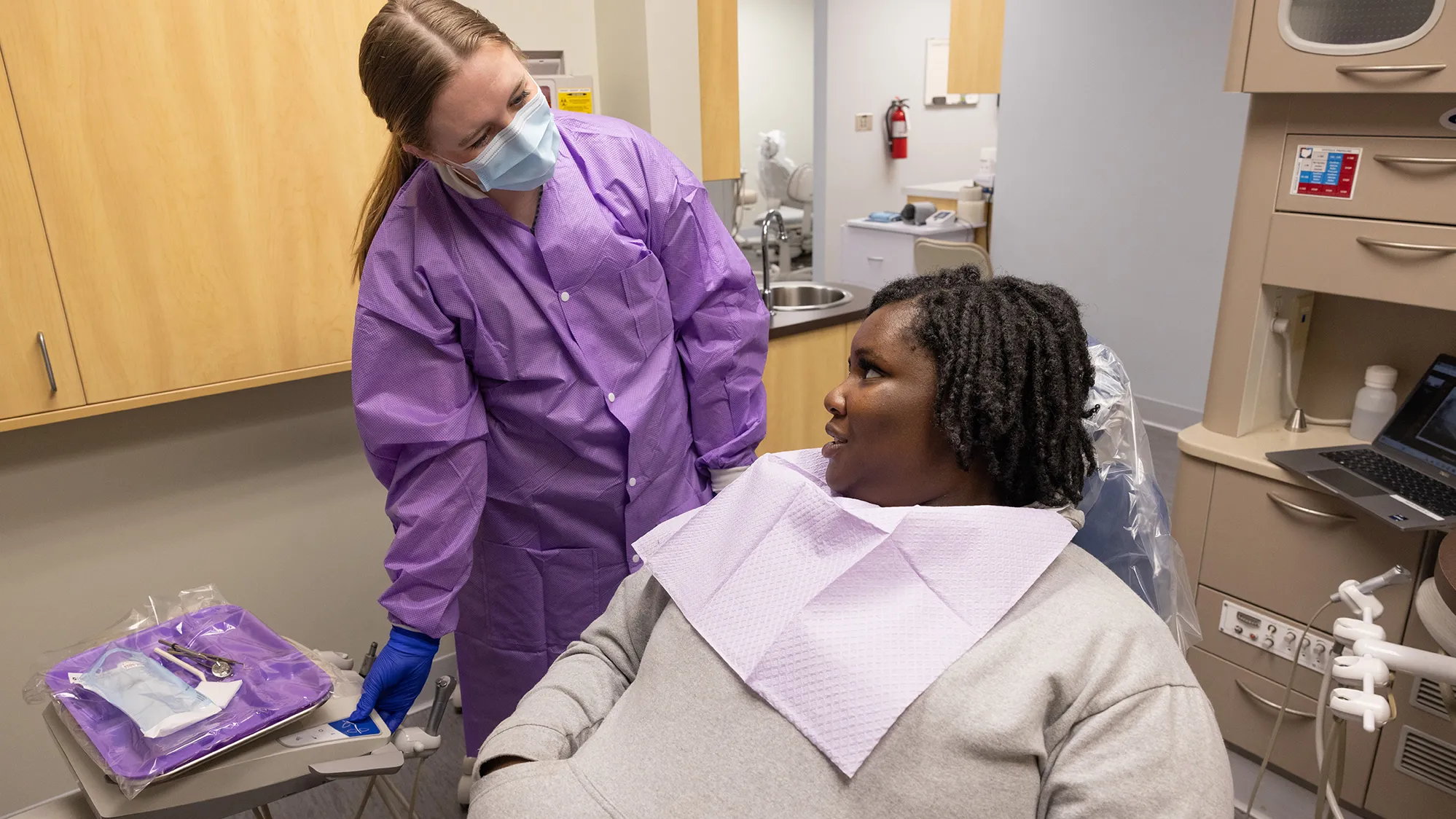 The student dentist, in a buttoned up scrub-style coat, leans sideways as she gives the young black woman in the exam chair her full attention. The student dentist wears a mask, but you can tell she’s smiling. The patient has the usual paper dental bib hanging around her neck and she’s looking directly at the student dentist.