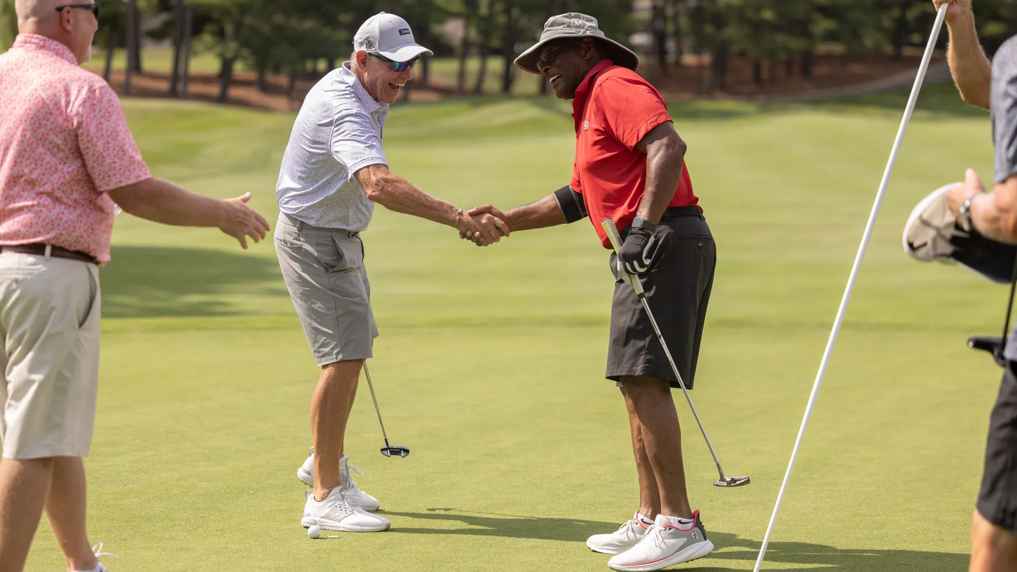 On the golf course, Archie shakes hands with a fellow player as another walks up to shake his hand, too. Archie’s wearing a wide brimmed hat and is smiling. Each man carries a golf club. 