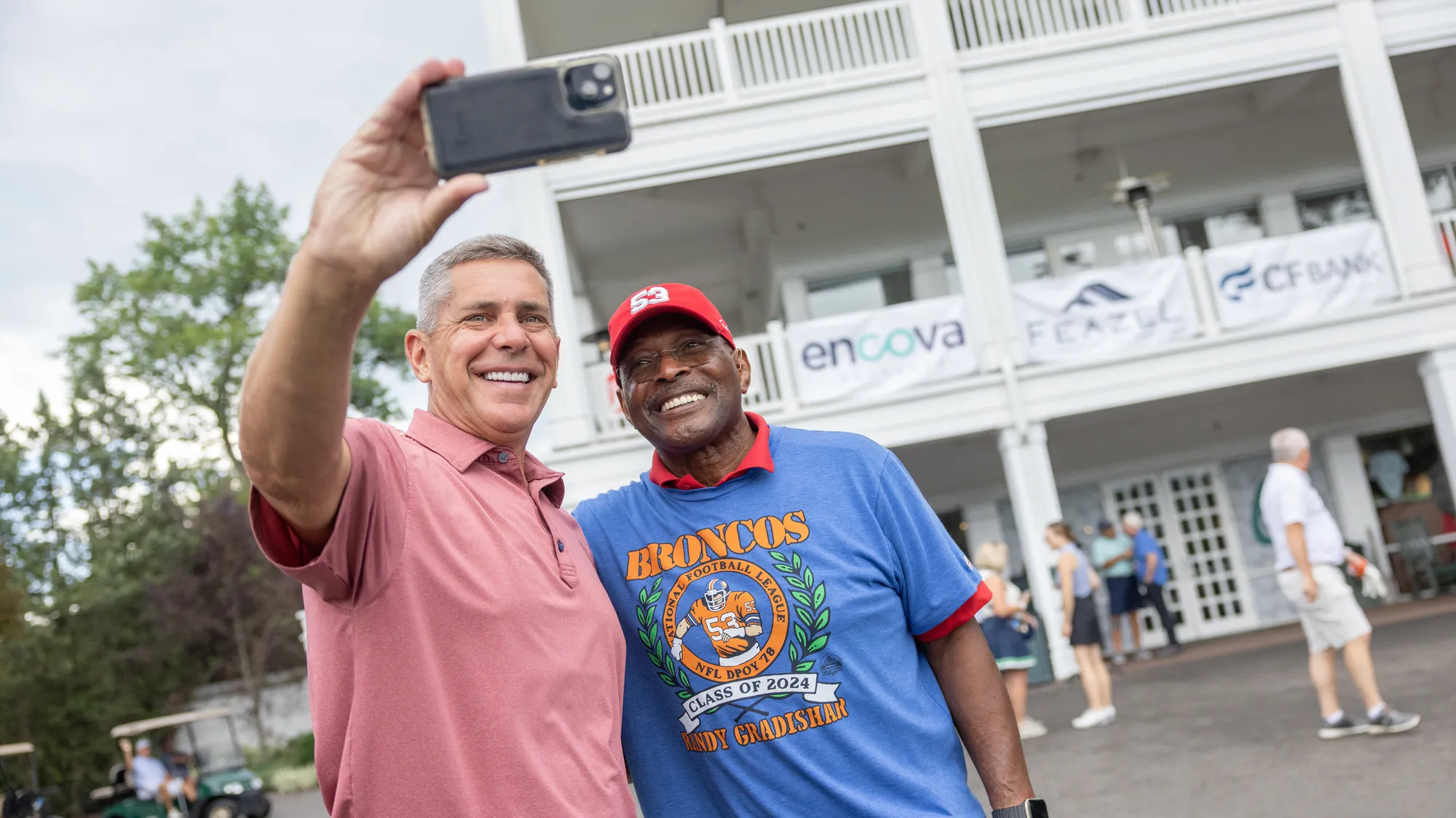 Archie Griffin grins as a man poses with him and hold up his cellphone to take a selfie. Both are in golfing clothes—Archie has a T-shirt honoring Randy Gradishar pulled over his golf shirt. They’re in front of a stately building at least three stories tall with a wraparound veranda on each floor. People mill about in the background. 