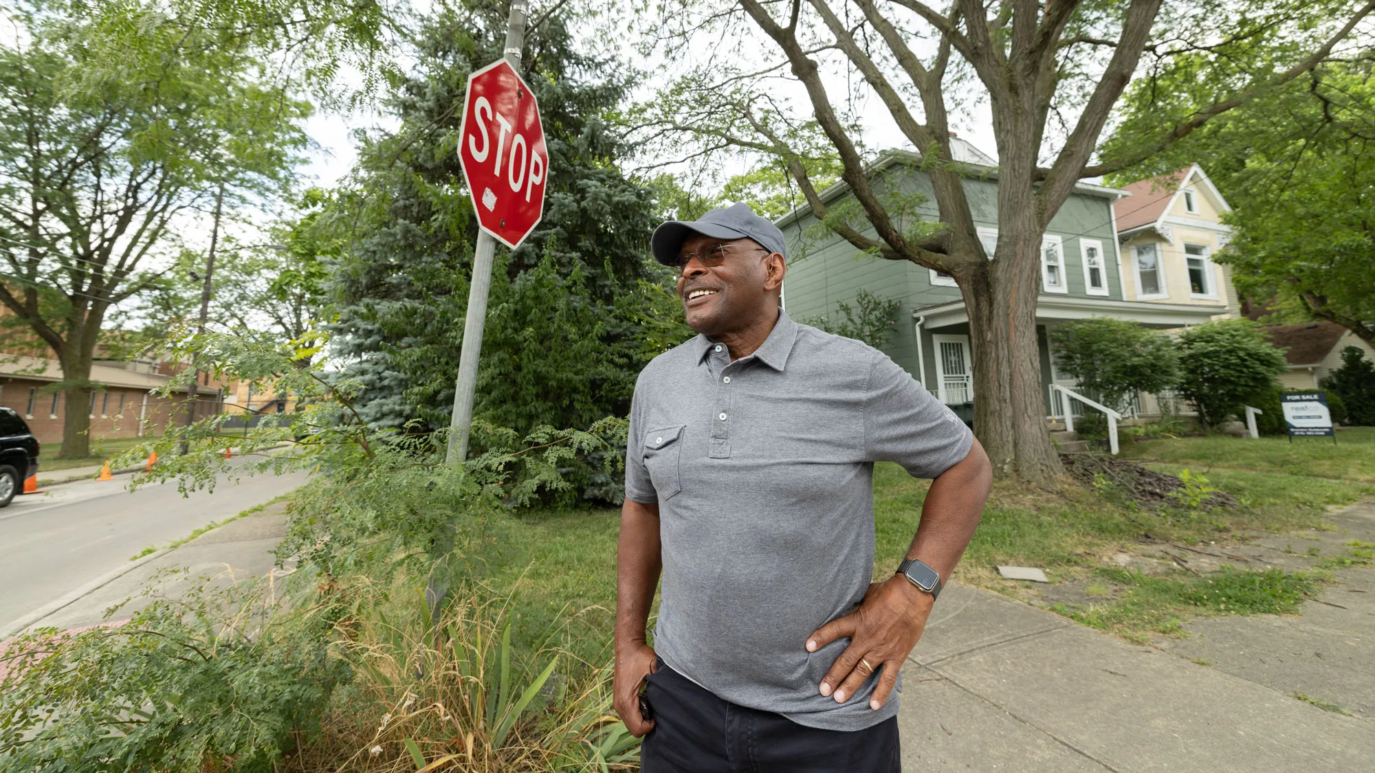 Archie Griffin smiles as he looks into the distance as he stands on the corner of two streets in a neighborhood. Next to him are out of season flowering plants and huge trees stand tall behind him. The houses appear to be the kind built in the early 1900s. Archie is wearing a golf shirt and a ballcap in the same color. 