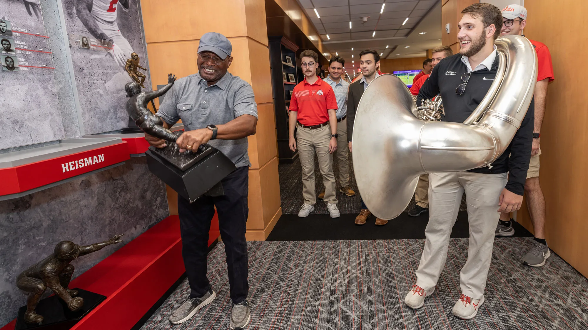 Archie Griffin hefts a replica of his Heisman Trophies—it must be heavy because you can see the muscles in his forearms. Watching are members of the marching band. The student in the front, a young white man with a beard, has a sousaphone wrapped around him. This was the day they invited Griffin to dot the i at a game this season. 