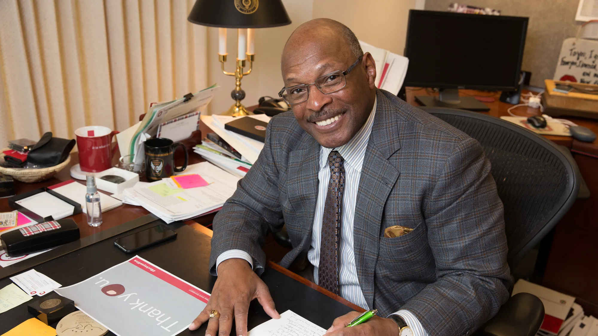 Archie looks up and smiles while sitting at his work desk writing a card. He’s wearing a plaid suit, striped shirt and patterned tie, and the desk behind him is piled with papers, many marked with sticky notes. 