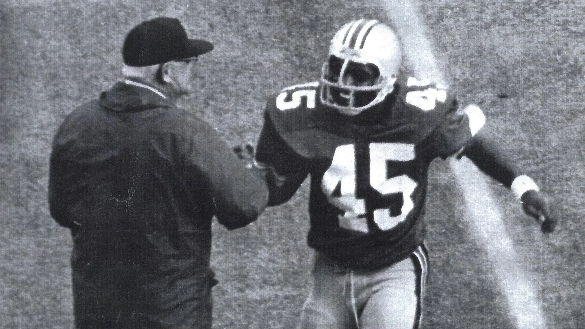 This historical photo taken from above, perhaps the first row of the stands, shows Archie Griffin as a college football player running toward the sideline. Under his helmet, he’s grinning as he clasps hands with Coach Woody Hayes, an older white man wearing a winter coat and ballcap. He’s mostly turned away from the camera. 