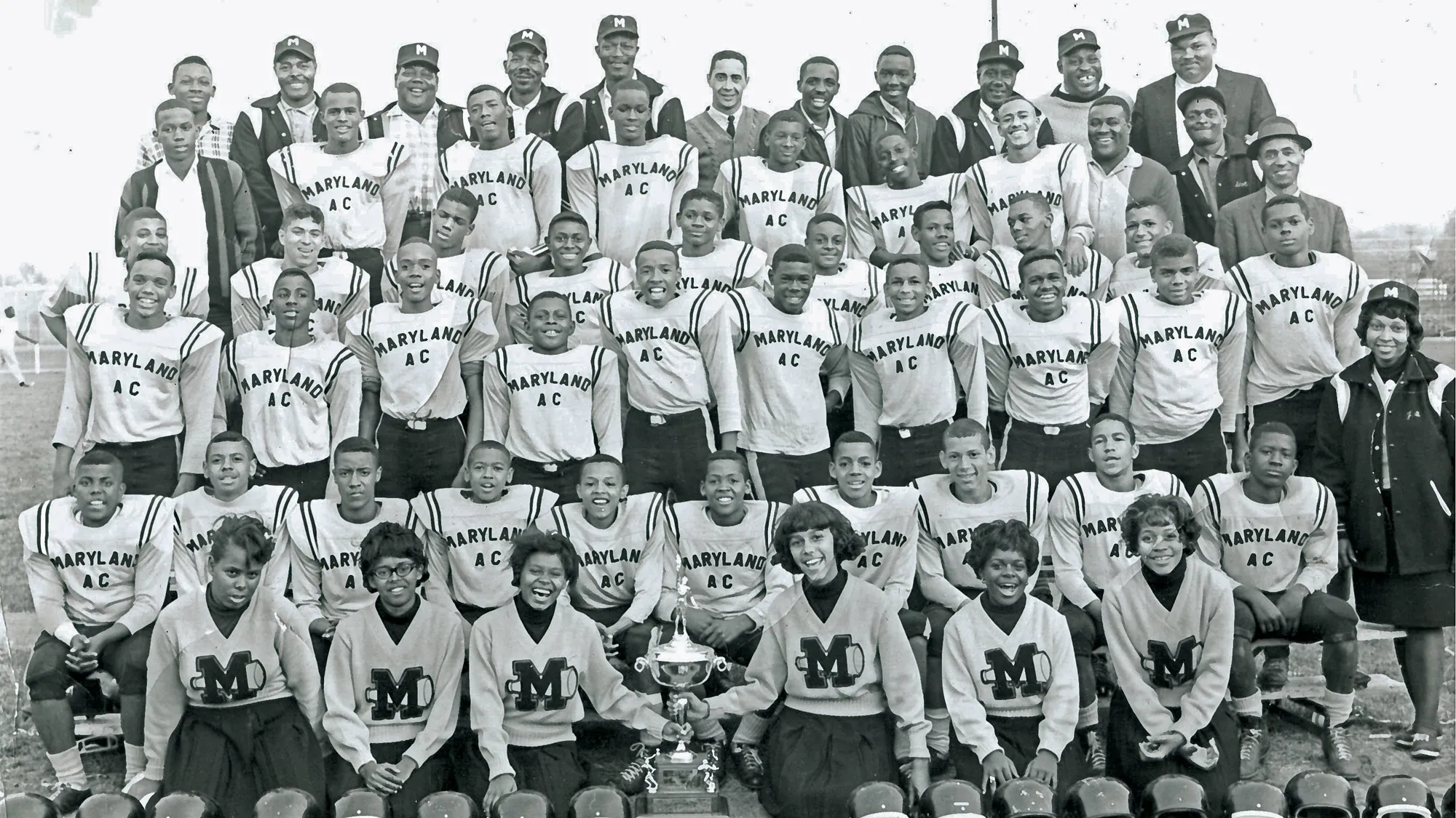 In the 1960s, a football team of 35 Black boys, 15 coaches, as well as six cheerleaders and their coach, pose for a team photo in 6 rows. A big trophy sits on the ground in front of them. Archie Griffin stands in the third row with a proud smile. He’s significantly shorter than the other in his row, standing about shoulder height. 