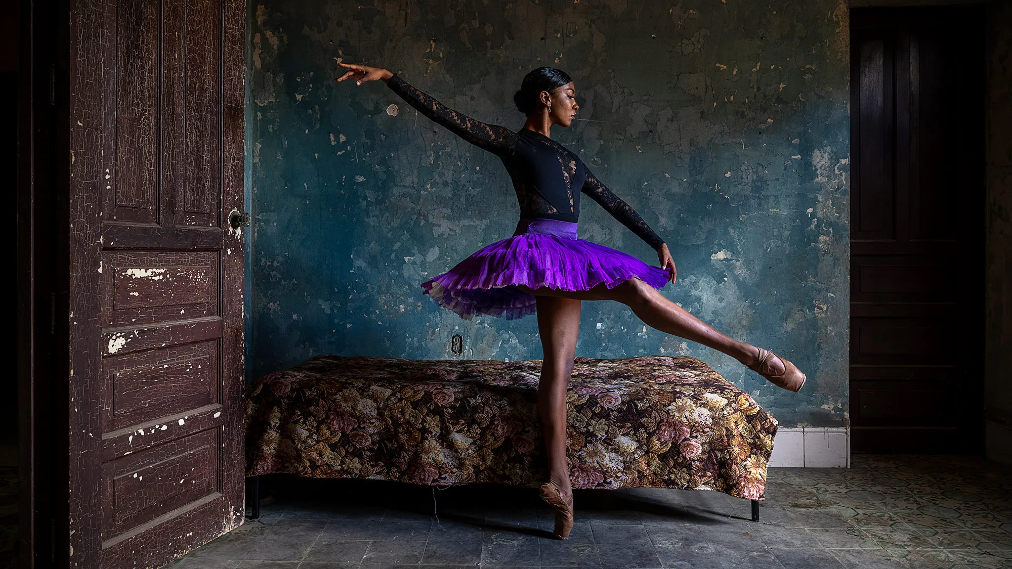 An elegant ballerina dances in a rundown bedroom with peeling paint and a flowered bedspread over a short bed. The dancer poses on one toe shoe with her other leg extended from her hip in front of her. She stretches her arms in opposite directions, parallel to her lifted leg. Her tutu shines, marking a bold contrast to her surroundings. Her facial expression is calmly determined. 