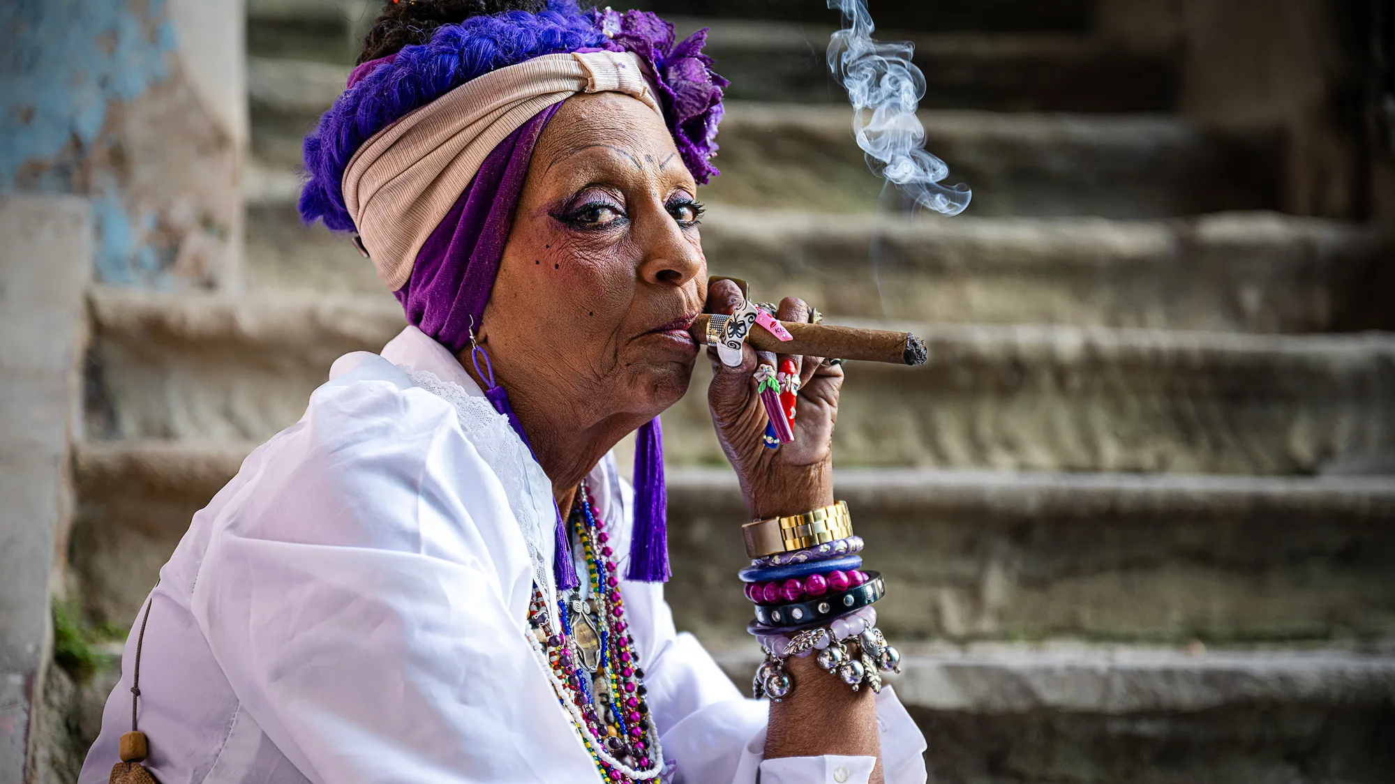 An older woman wearing a crisp white shirt, lots of beaded necklaces and bracelets, a multicolored head wrap and flapper-style makeup sits on stairs while smoking a cigar. 