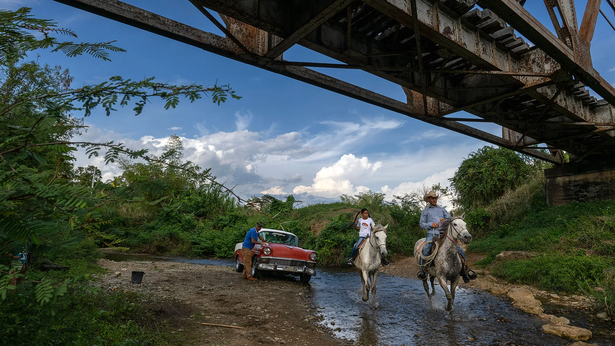 An old-fashioned Chevy suts on the dirt bank of a narrow river as a man polishes one round headlight. Closer to the camera, an adult and child ride matching white horses down the middle of the waterway. Overhead is an old steel and wooden bridge. The river curves out of sight at the far edge of the photo. The sky is bright blue and the clouds are wispy. It’s a charming scene. 