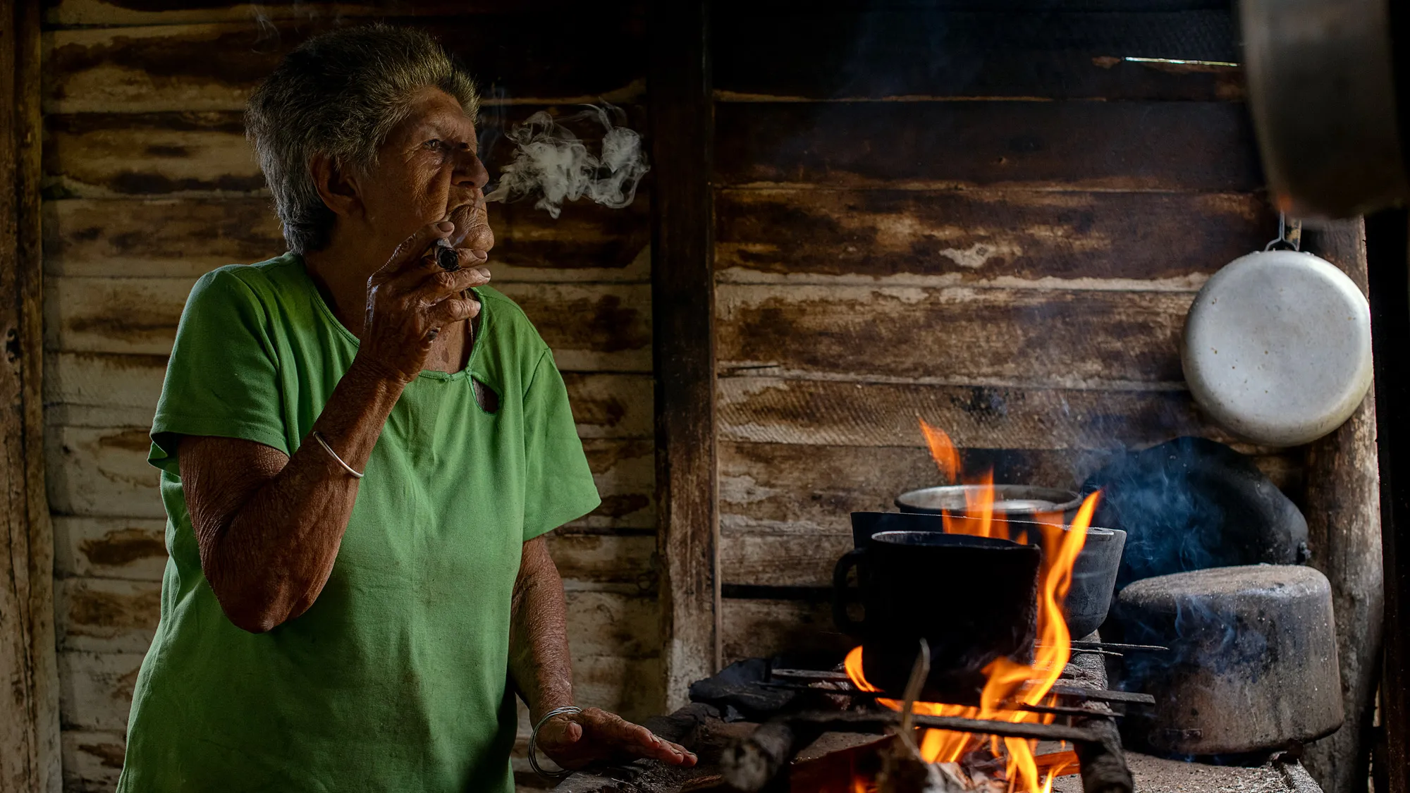 An old woman in an oversize T-shirt stands over a stove smoking a fat cigar. The stove has a pile of wood on top that has been lit on fire to heat a cast iron pot. The walls behind her are weathered wood planks. 