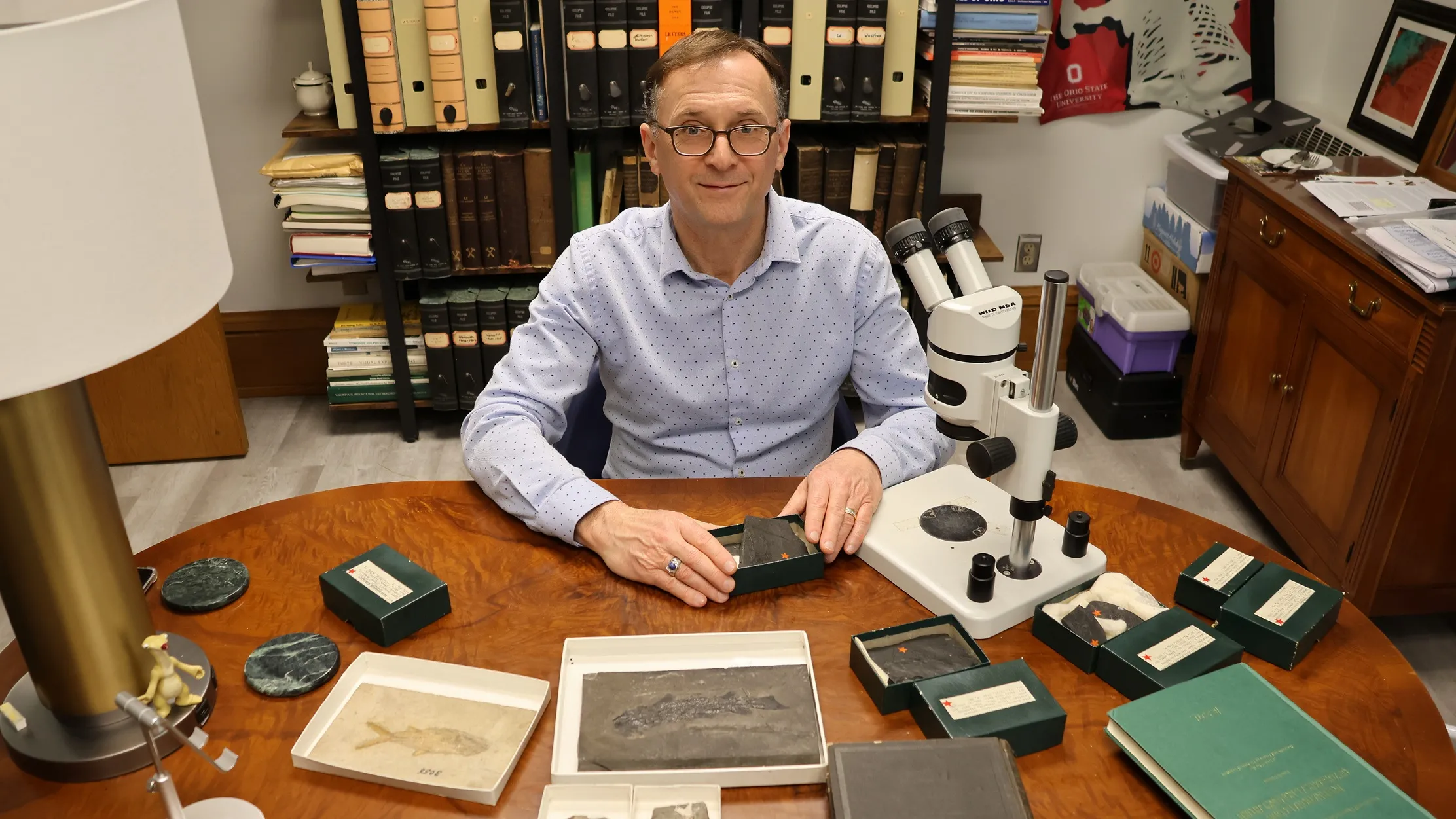 Loren Babcock, a thin white man with glasses, gray hair at his temples and a stylish button down shirt, shows a set of fossils of various sizes, each in its own shallow box. They’re flat rocks with fish impressions that show as shinier or darker. Also on the table is a microscope. Baback looks pleased and like he’s getting ready to share a fascinating story about the one he holds. 