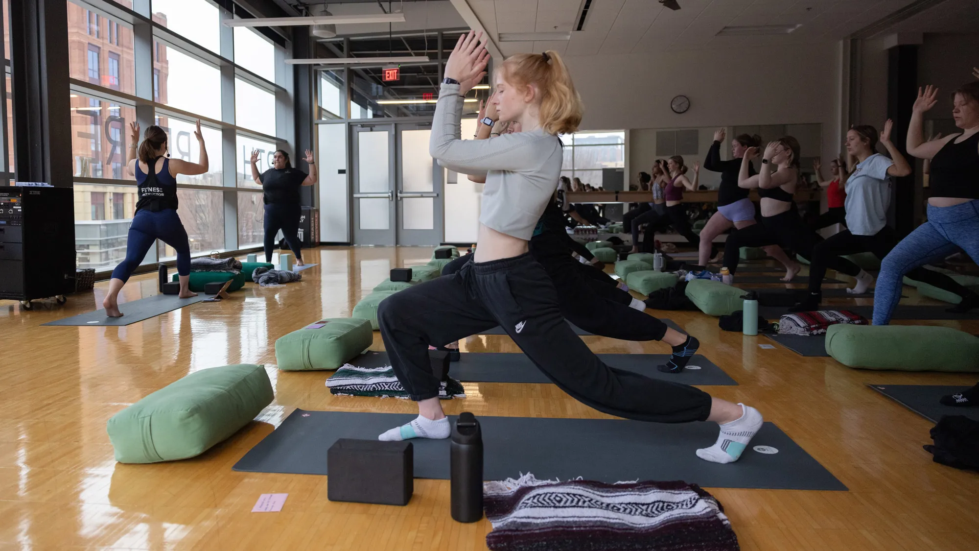 Lines of Buckeye students, each on their own yoga matte, strike the yoga pose shown by the teachers. It is right leg forward and bent so the flat of the foot is on the floor; left leg stretched far behind, so only the ball of the foot is on the floor; and elbows bent 90 degrees and hands held just above head height.