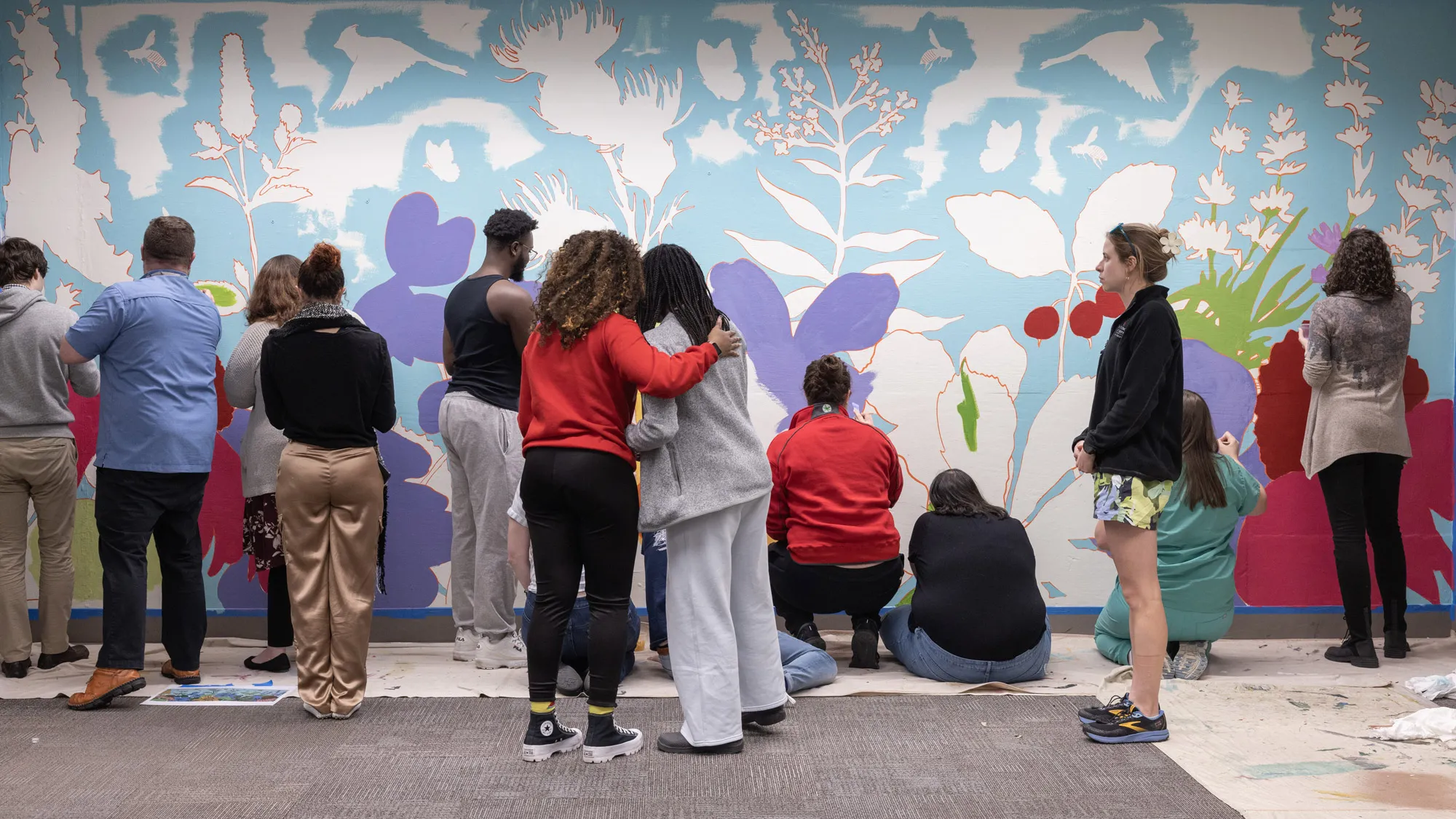 More than a dozen students face away from the camera as they work on painting a colorful wall of flowers and leaves. Birds and flying insects can also be seen outlined against the color used for the sky. The students are a range of sexes, races and clothing styles.