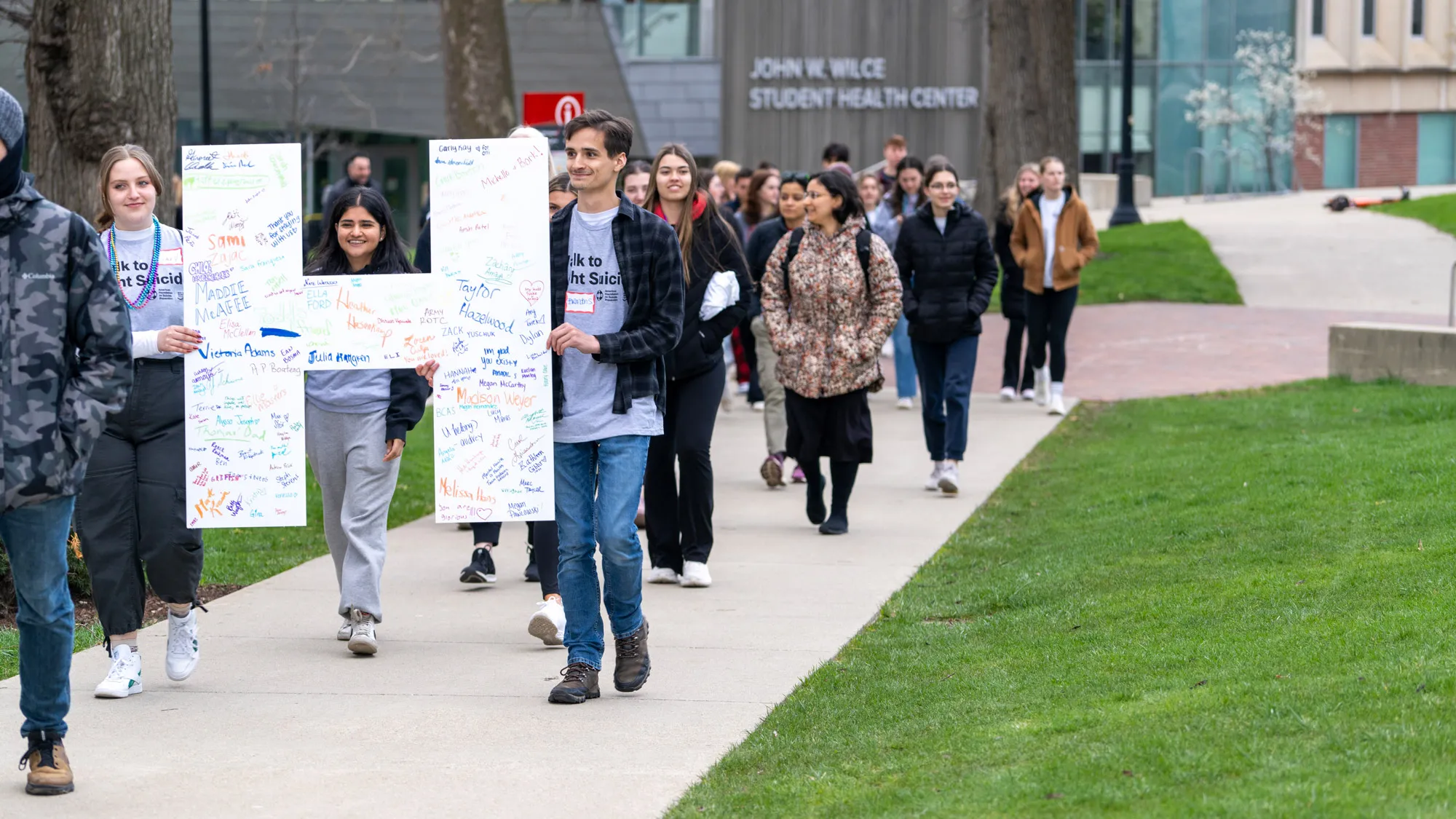 A line of people walk up a sidewalk; in the background is the John W. Wilce Student Health Center. The people carry a large H with handwritten messages in marker on it. Many wear coats but a few of their T-shirts can be seen. They say “Walk to prevent suicide.” About 25 people are shown in the photo, but they are the back end of the procession.