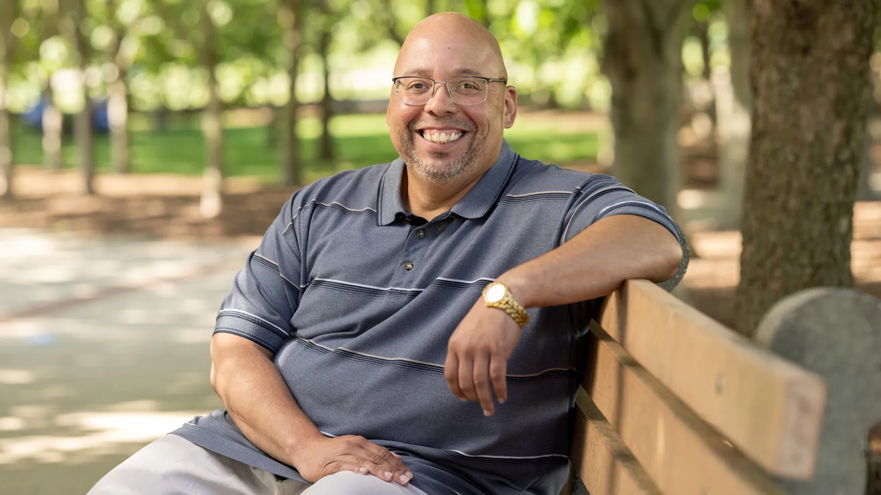 Shawn Lucas, a white man with shaved head and glasses, sits on a park bench with one arm propped along the back. His smile seems both genuine and inviting—he looks like the kind of man students could easily feel at ease with.