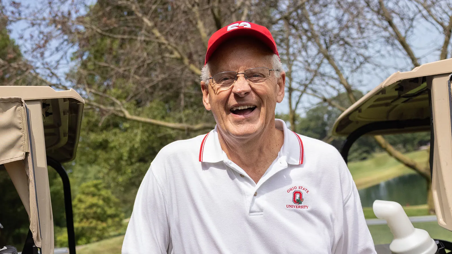 Randy Gradishar, an older white man wearing golfing clothes and a ballcap, slightly leans on a golf cart as he laughs during a conversation with someone not shown in the photo.  