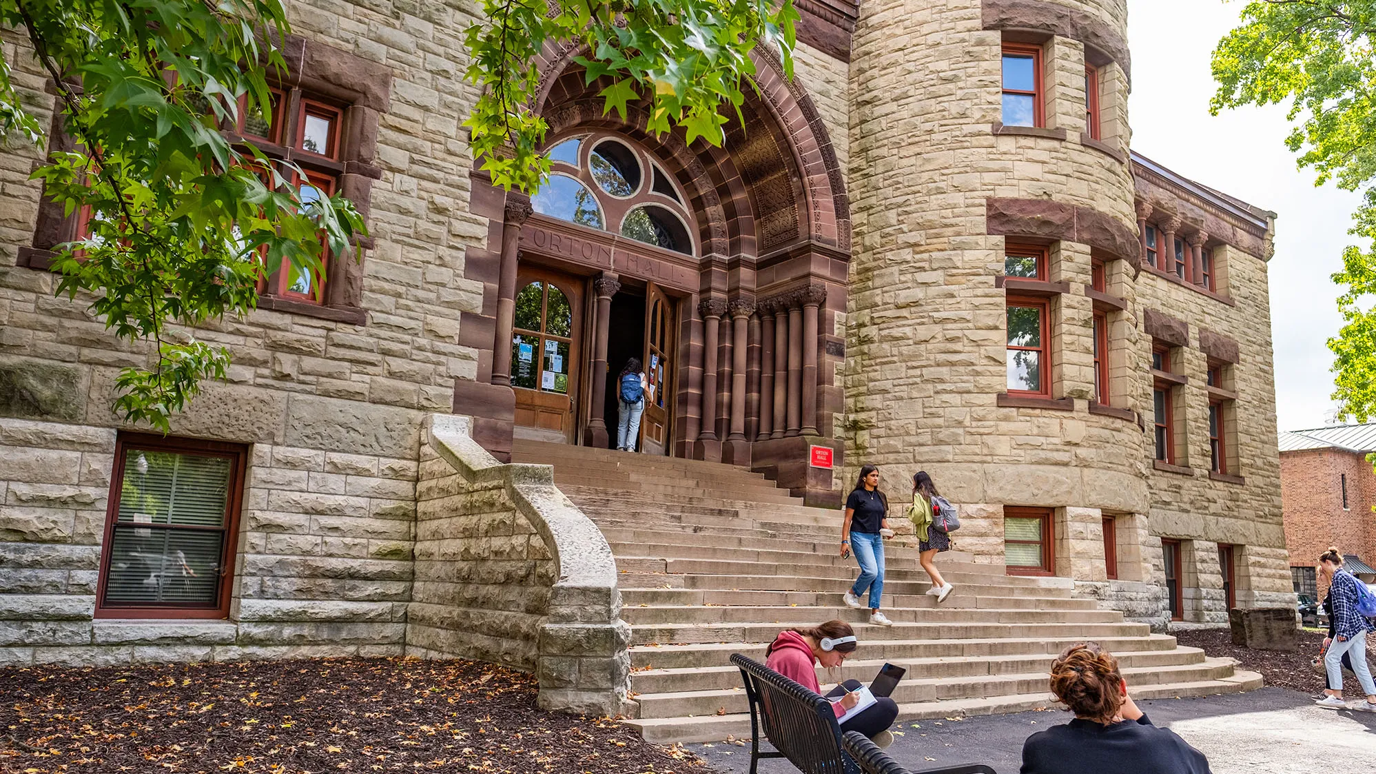 Orton hall is a stone building with 20 wide stone steps leading to the arched main entrance. The stone work is special and represents the different geological period of earth. Students walk in and out in this photo, which shows the stateliness of the building on a pretty day.