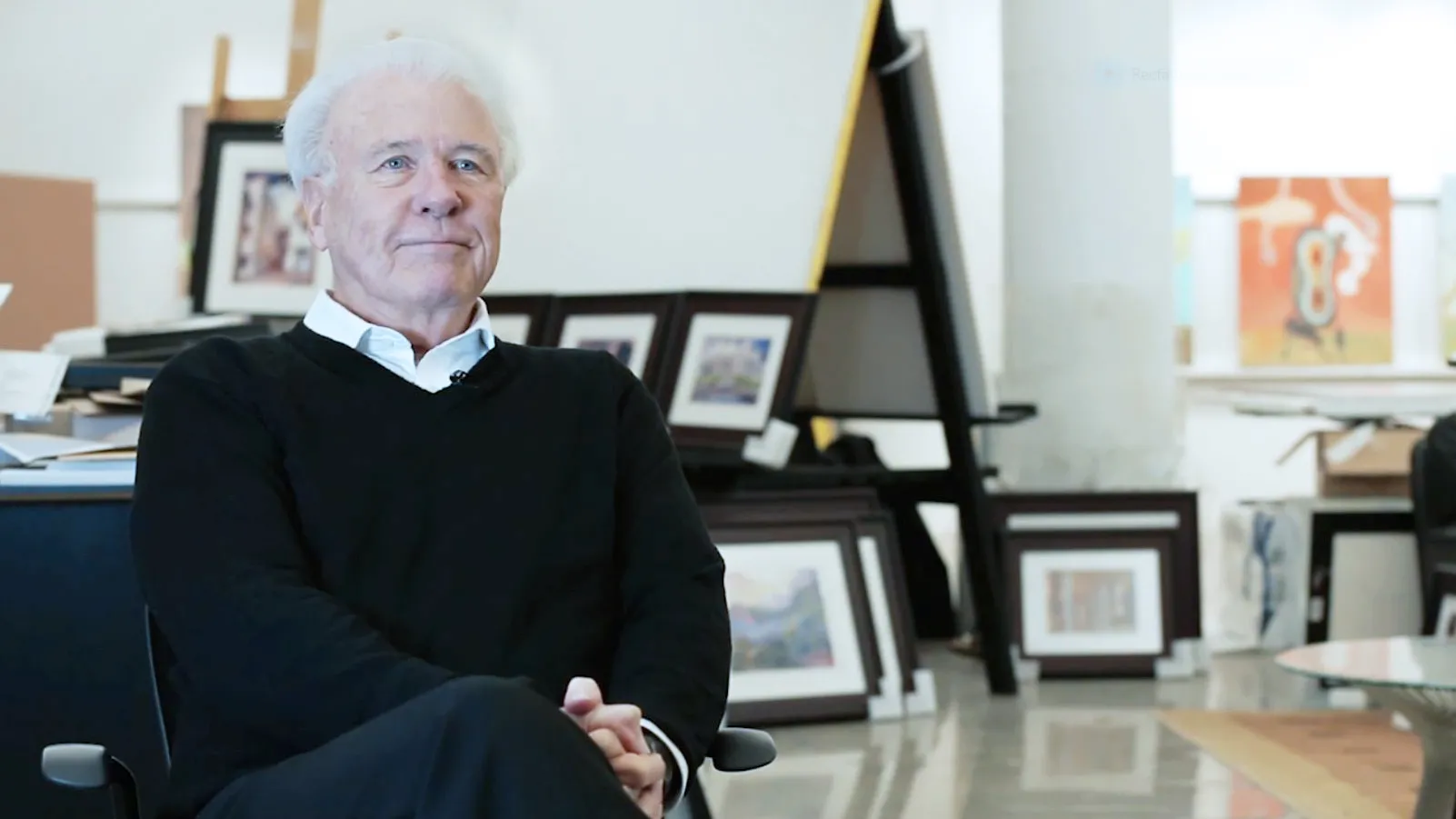 George Acock, an older white man with pure white hair, sits with hands clasped and legs crossed, looking happy, confident and comfortable in a studio where canvasses line the wall and other available space.