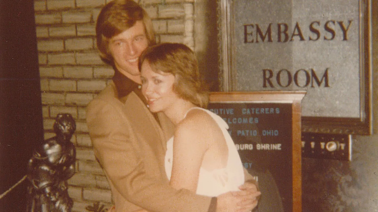 In this old photo, a college-age couple hugs as they pose, clearly comfortable being affectionate with each other. They’re both dressed up and have the feathery hair of the 1970s.