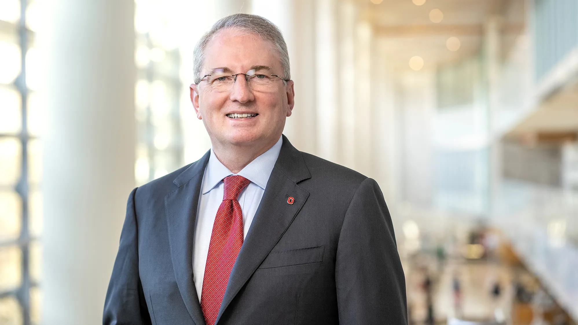 Dr. John J. Warner looks friendly and smart as he poses for a photo in a grand space where pillars and windows line a wall and study tables can be seen behind him. Warner, who is smiling, is a middle age white man in a suit with glasses and a dimple.