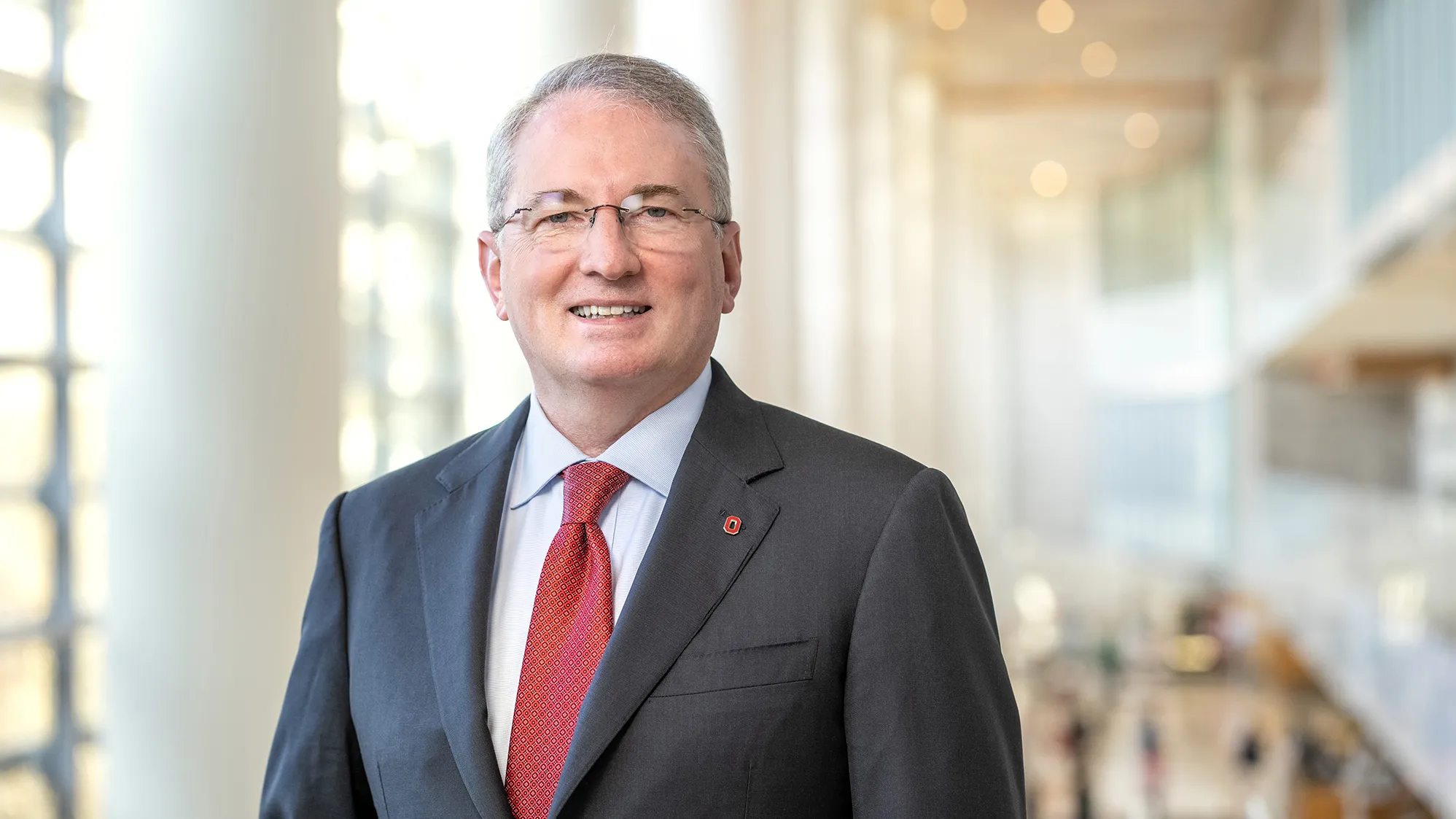 Dr. John J. Warner looks friendly and smart as he poses for a photo in a grand space where pillars and windows line a wall and study tables can be seen behind him. Warner, who is smiling, is a middle age white man in a suit with glasses and a dimple.