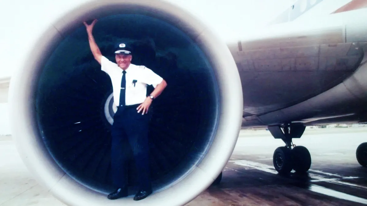 In this old photo, David Harris poses for a snapshot while standing on the edge of one of the engines on a jetliner. It's not powered on, and the full plane can't be seen. Harris looks like he's having fun as he reaches one arm up to touch the top of the round engine holder.