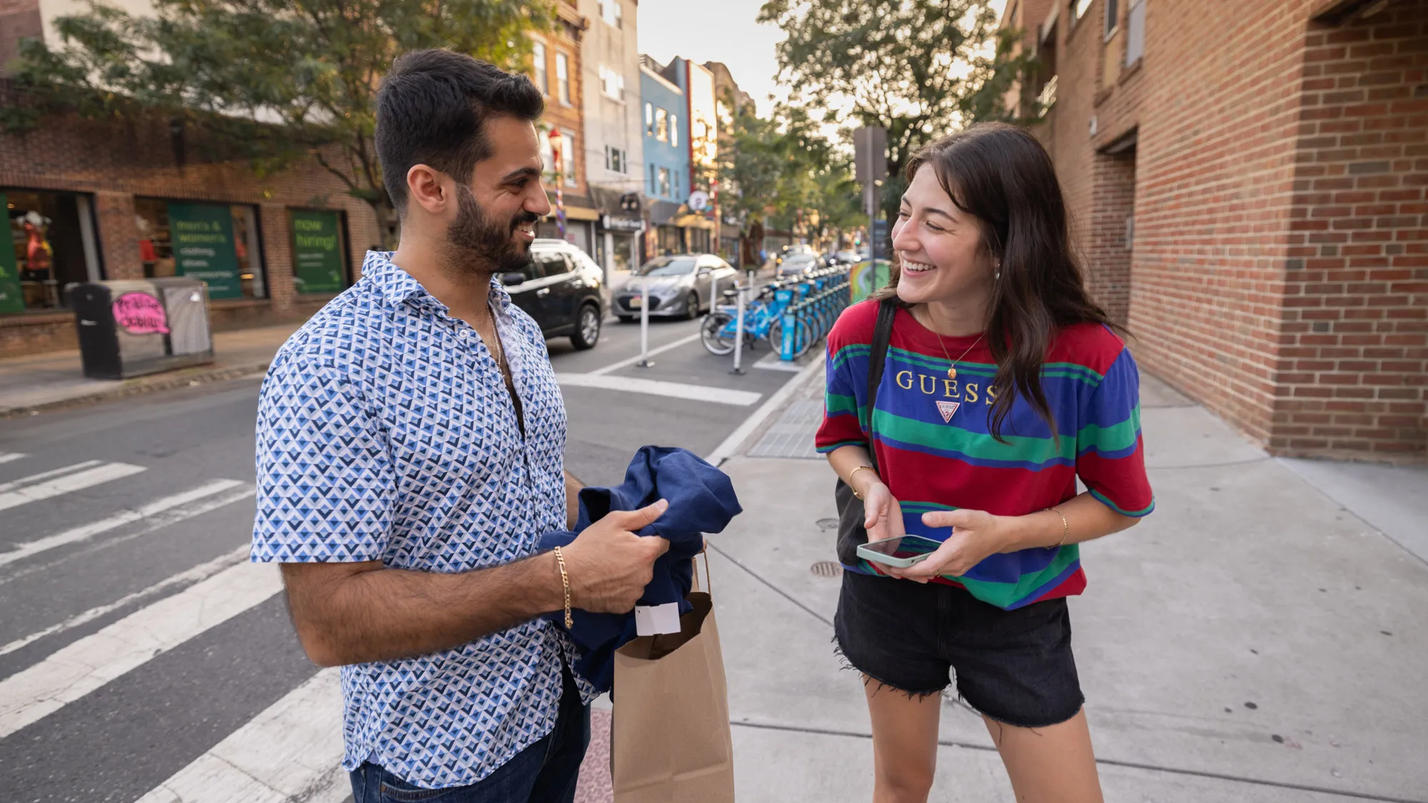 Tovah Kaiser laughs as she talks with a friend on a Philadelphia sidewalk. She’s wearing a Guess-brand striped T-shirt and shorts and leans forward as she laughs. Her friend has on a geometric-patterned short-sleeve button down and holds a piece of clothing he’s pulling out of a brown paper gift bag. He has a beard and looks to be of Middle Eastern descent. 