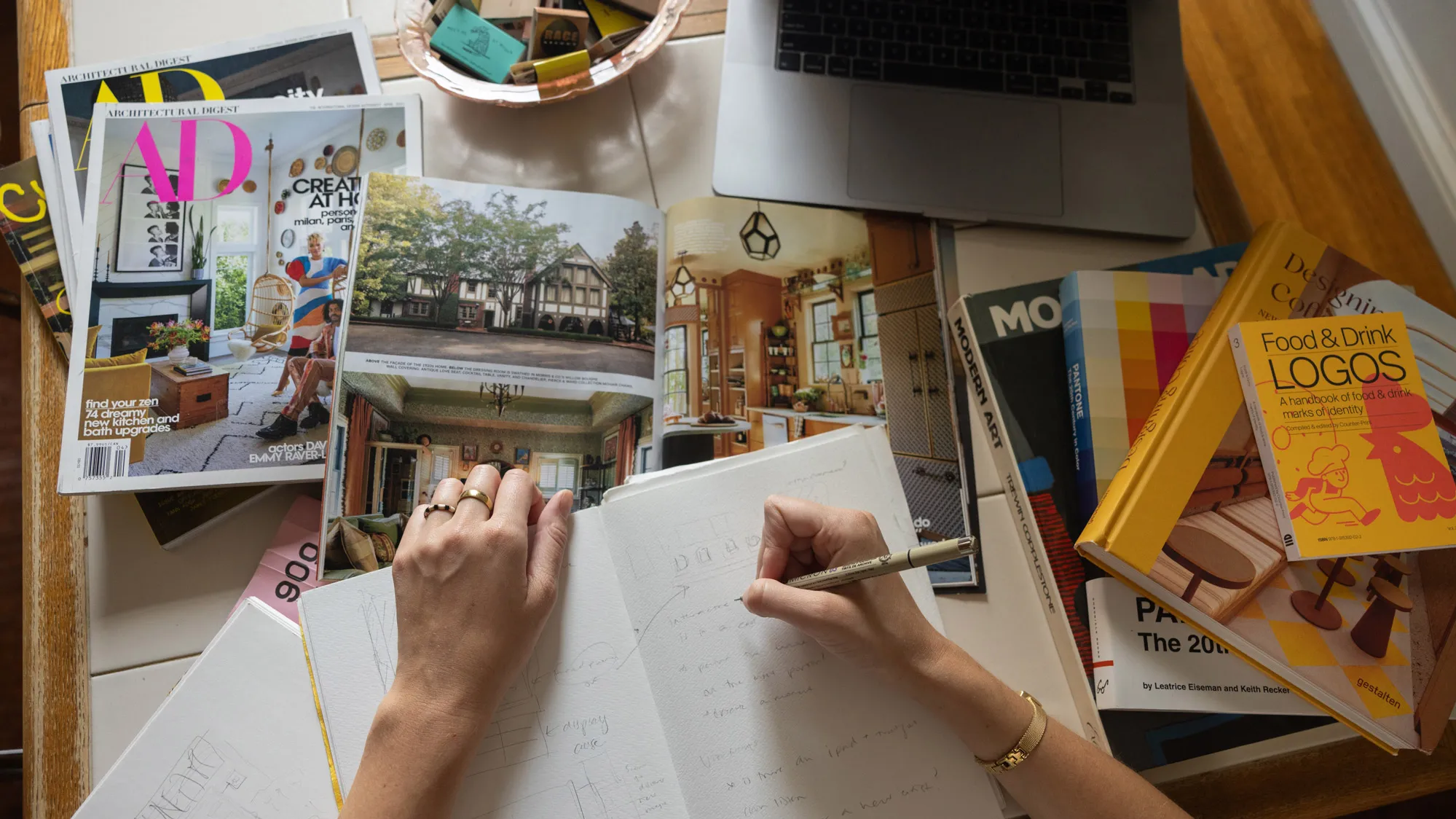 This photo shows a hand writing in a notebook with unlined pages. Tovah has written sentences and sketched some pictures in pencil—they cannot be made out. She has the notebook sitting on colorful magazines, such as Architectural Digest, and next to the notebook is a stack of design books. Words on the top that can be made out include “Food & Drink” and “logos.” 