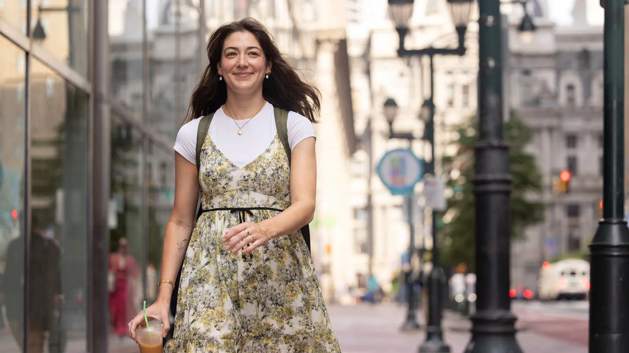 Tovah Kaiser smiles as she strides down a Philadelphia sidewalk. She’s a young white woman with long hair and a tattoo of a simplified fish on the inside of her right arm. She’s wearing a pretty and floaty floral dress over a plain white T-shirt along with gold earrings, necklace, bracelet and rings.