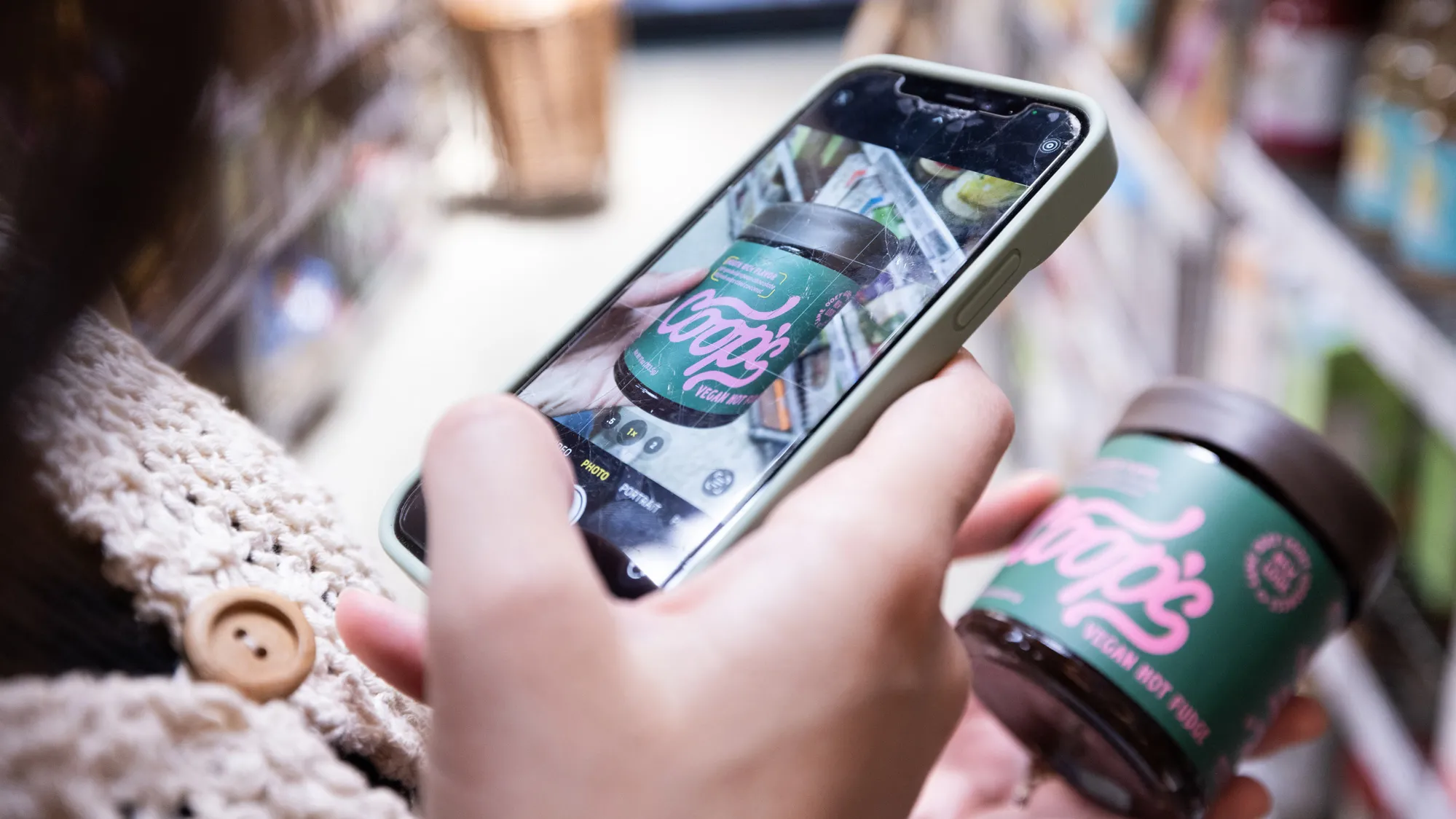 This photo shows a hand holding a squat jar of fudge sauce. The label is dark green with a pink font that says “Coop’s” in cursive and in print: Vegan nut fudge” and “smooth rich flavor.” 