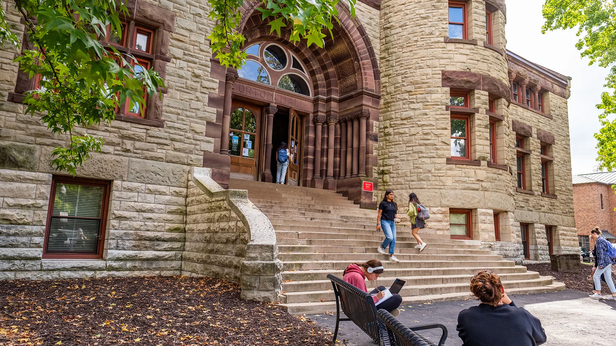 Orton hall is a stone building with 20 wide stone steps leading to the arched main entrance. The stone work is special and represents the different geological period of earth. Students walk in and out in this photo, which shows the stateliness of the building on a pretty day.