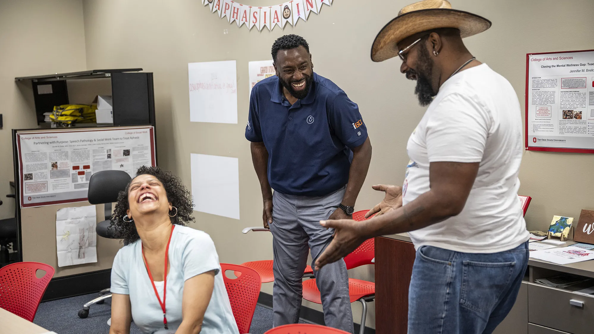 Three people laugh as they talk in a room with flags spelling OSU Aphasia Initiative on the wall. Two of them are husband and wife. She has her head thrown back as she laughs hard; her hoop earrings swing with her movement. Her husband, in dress pants and golf shirt, laughs with his hands on his thighs as he watches the man entertaining them with his storytelling. That man wears a cowboy hat and glasses. All three are black and middle-aged.