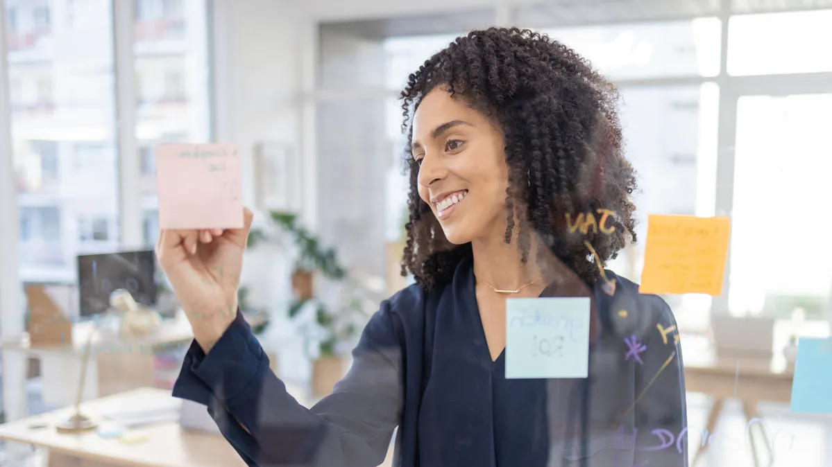 A young woman of mixed race smiles as she writes on a sticky note attached to a see-through project space in an office setting. The notes could be used for planning or leaving encouraging messages for co-workers; the text can't be read in the photo.