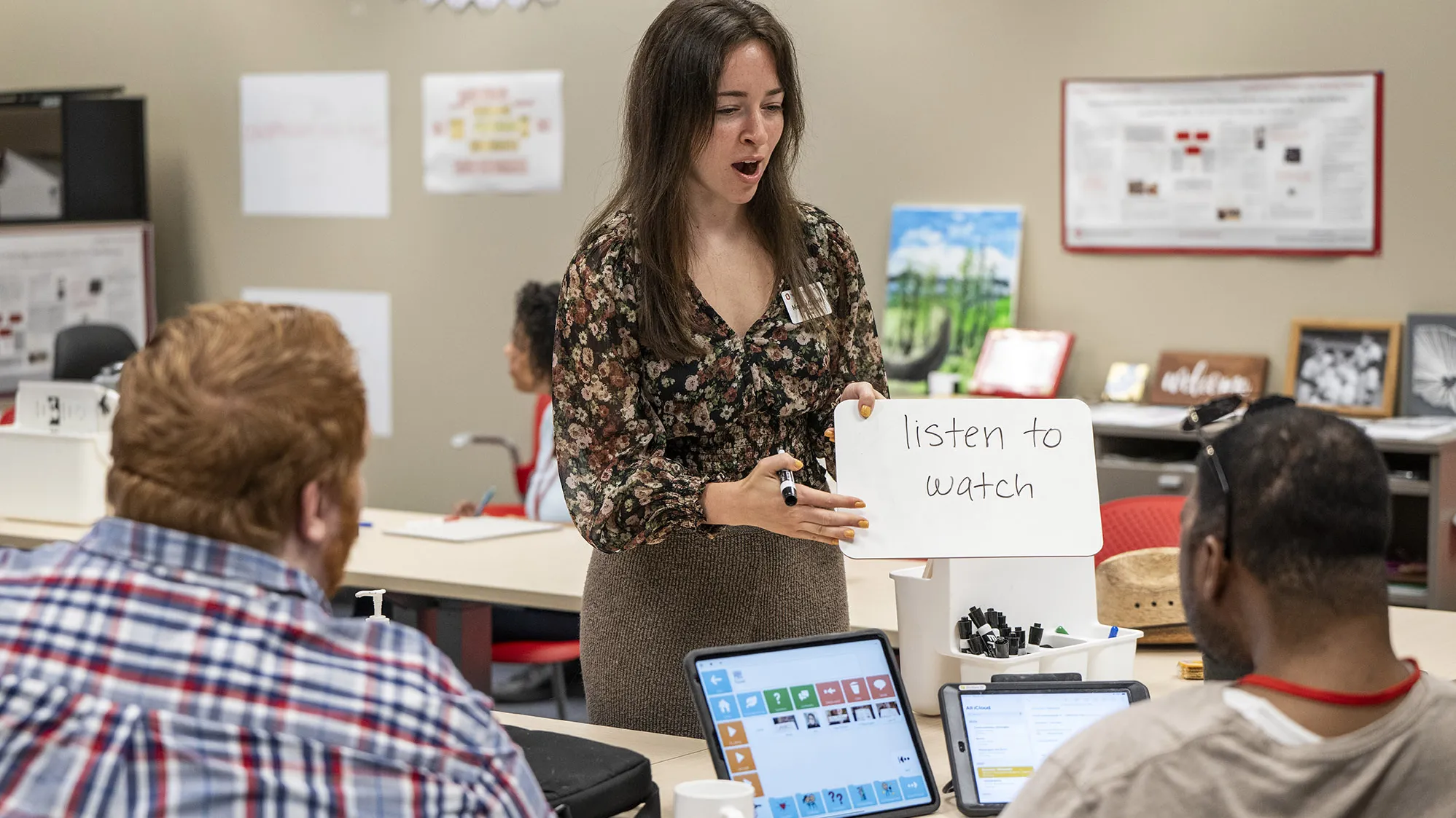 As she walks along the inside of the U-organized worktables, Monica O’Neill, a young white woman with long hair, stops in front of one participant, who is seen only from behind. She holds a small marker board that says “listen to watch” and her mouth is open as she enunciates the words.