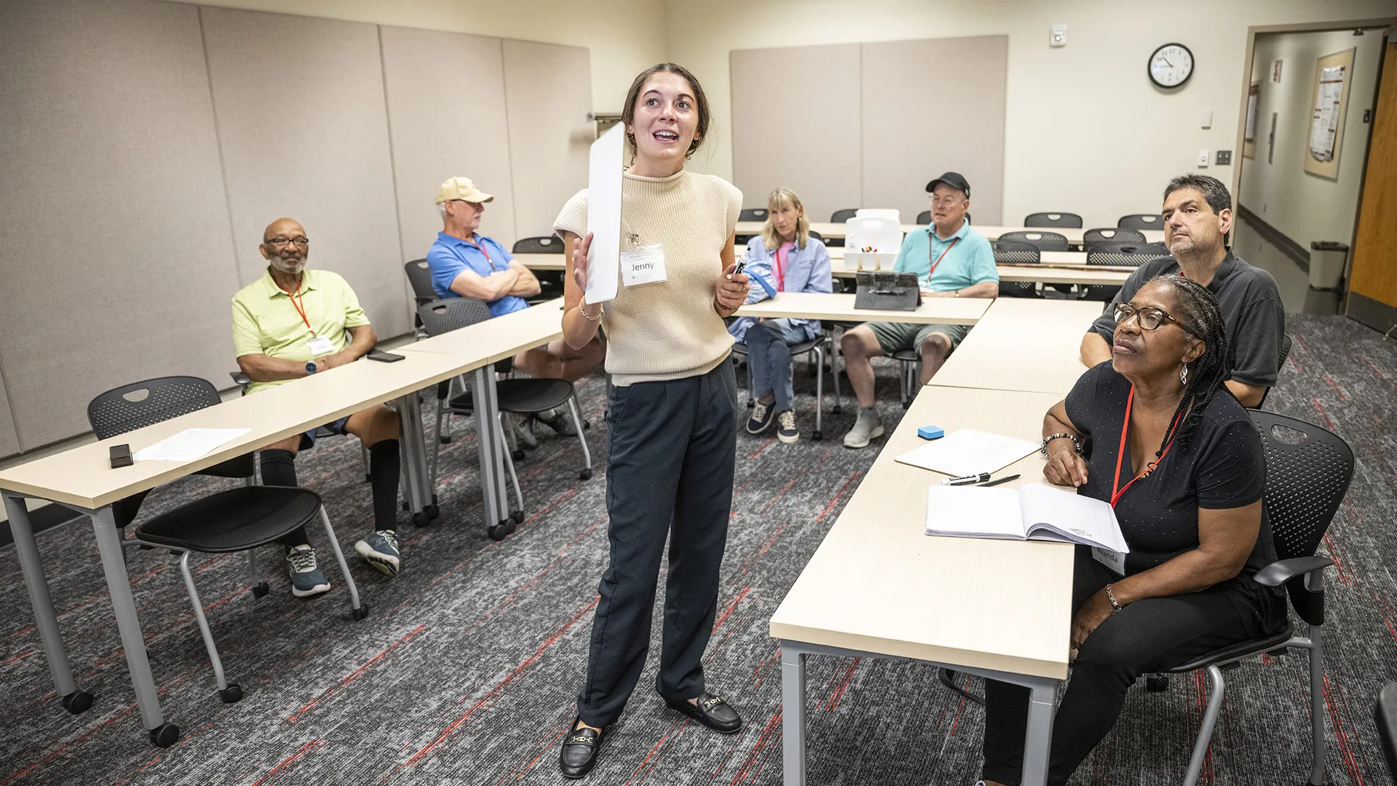 A young white woman stands in the middle or worktables pulled into a U shape. She and the people who sit at those tables are all looking out of the frame of the photo, at a screen on the wall. There are at least six people at the tables, a mix of races, ages and genders. 