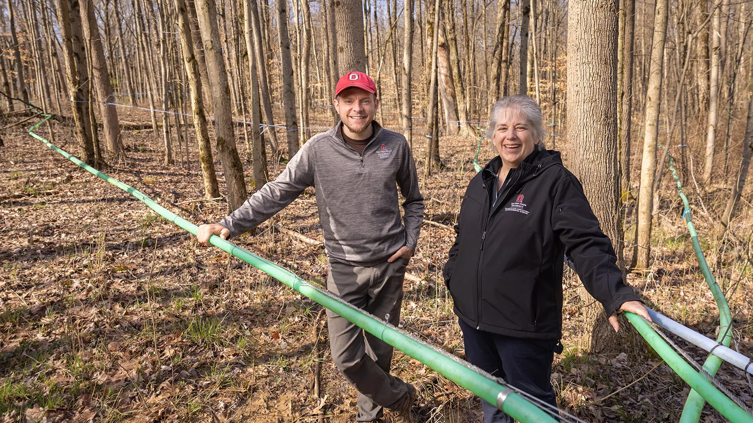 Two people wearing Ohio State-branded sweatshirts stand in woods during winter. Green tubes weave between the trees and around the people, who are laughing as they look at the camera. Hints of bright green grass peaking through the forest floor suggest spring is in the air.