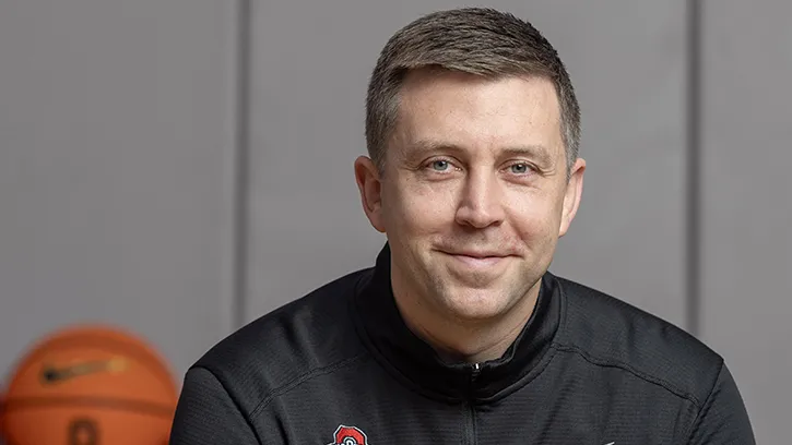 Ohio State Basketball Coach Jake Diebler holds a basketball while sitting on a stool and smiling warmly at the camera. He looks like he is about to laugh. Diebler is a white man who looks to be in his 30s. He has short cropped hair, blue eyes and wears black workout gear.