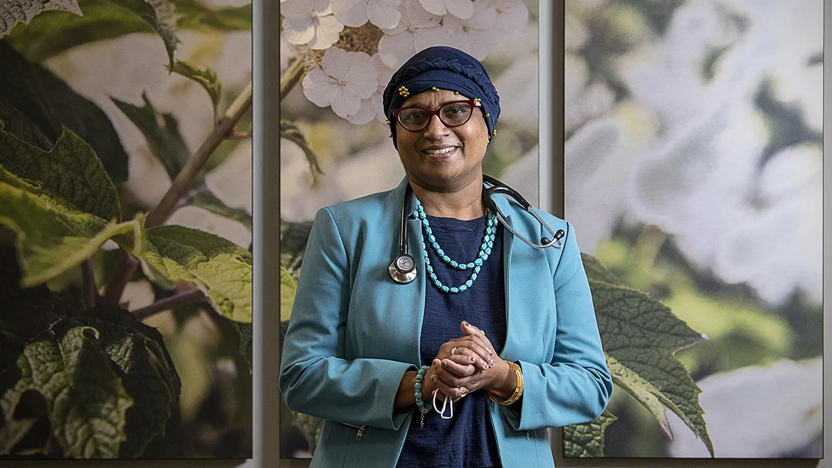 Dr. Bhuvaneswari Ramaswamy wears a headscarf wrapped around her head and glasses as she smiles for a portrait. At this time, she was undergoing cancer treatment and caring for patients. She looks liek someone you'd want to trust. A middle-age woman born in India, she wears a business suit, a stethoscope around her neck and turquoise necklace and bracelet. The background is a wall with a huge photograph of flowers.