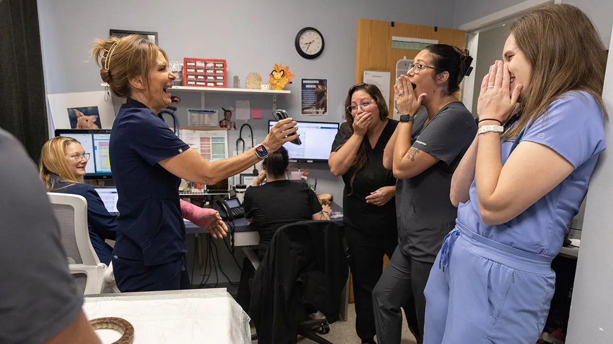 In a workroom at the veterinary office, Dr. Oglesbee holds out her phone while three women standing and looking at it seem to gasp. They have huge grins and their hands in front of their mouths—they’re all full-body laughing. Two more people in seats in the background also seem to laugh.