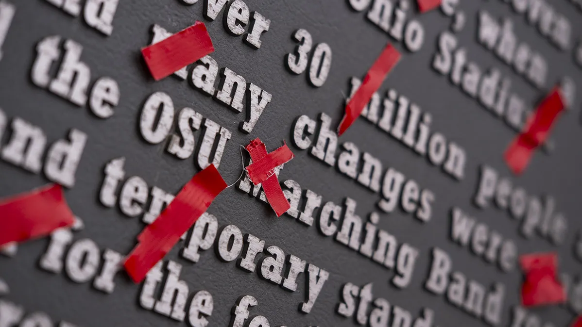 A gray plaque outside ohio stadium has every single m crossed out with bright scarlet tape.