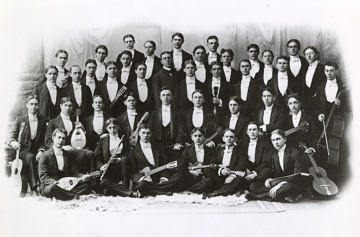 In a very old black and white photograph, about 40 men, each wearing a tuxedo, pose for a photograph. Many hold stringed instruments and their expressions are mostly serious.