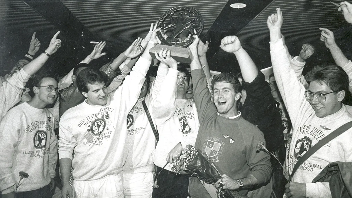 About a dozen members of the men's glee club hoist a trophy as they cheer. The photo is black and white, giving it a dtaed look, even though color photography was available at the time.