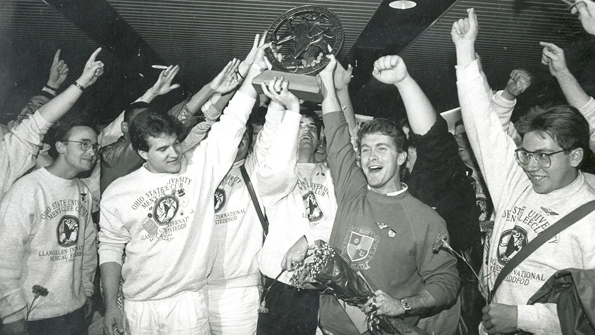 About a dozen members of the men's glee club hoist a trophy as they cheer. The photo is black and white, giving it a dtaed look, even though color photography was available at the time.