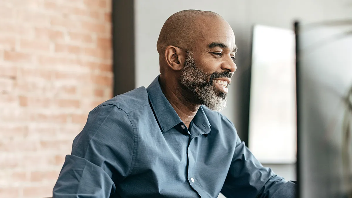 This image shows a Black man with a bright smile sitting at a desk and using a computer