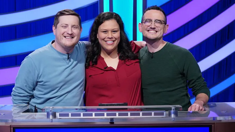 Three Buckeyes pose with their arms around one another on the stage of Jeopardy! The two on the sides are men; in the center is the woman. They seem genuinely close and pleased to be competing together.