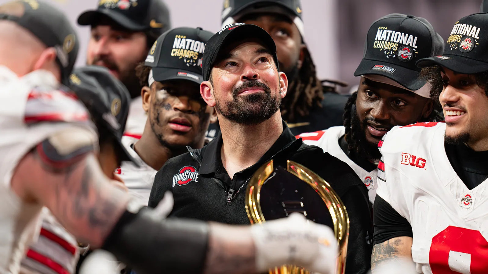 Ohio State football coach Ryan Day, surrounded by football players, looks proudly upward. He appears a little tearful but is smiling. Everyone in the photo taken after they won their final game of the playoffs wears a National Champs baseball cap.